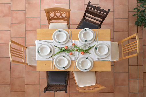 Wooden table setting with six empty white plates and bowls, cutlery and some tulip flowers, different vintage chairs around on a tiled terracotta floor, high angle view from above, selected focus