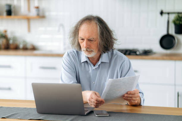 istock Disappointed upset middle aged gray haired caucasian man sits in the kitchen at a table with laptop, holds documents in his hands, does paperwork, reconciles data, analyzes the results of work 1495485918