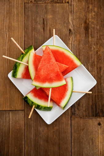 This is an outdoor photograph of sliced red see this watermelon on a white square modern plate sitting on a wooden picnic bench outdoors for a simple and concept of healthy eating and snacks during the summertime fun.