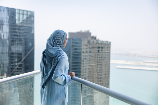 Mature middle eastern women is enjoying beautiful view from balcony. She is wearing blue dress. Focus on foreground.