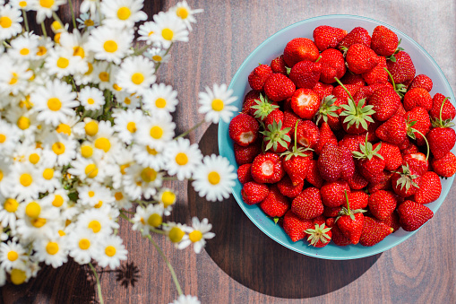 Ripe juicy strawberries in a bowl on the table, next to a bouquet of daisies in a vase. top view, flat lay