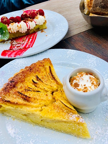 Vertical closeup photo of two desserts in a French restaurant: Pear and Almond Tart and Raspberry and Almond cake, both served on white porcelain plates on a wooden table at a French restaurant in Aix-en-Provence, south of France, in Spring.