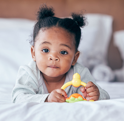 Cute, portrait and baby with a toy on her bed playing and relaxing in her modern nursery. Childhood, playful and African girl infant or newborn playing with plastic object in her bedroom at her home.