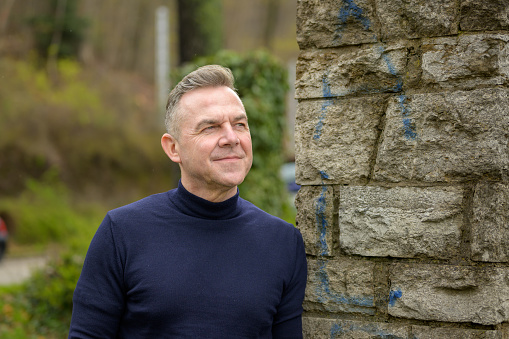 Middle age grey-haired attractive man wearing a blue sweater is standing near a wall of large stones and looking into the distance with a gentle smile