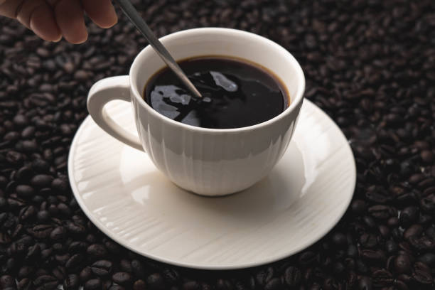 Hand holding coffee stirring spoon in white cup on table. Black coffee in white cup isolated on background of roasted coffee beans.