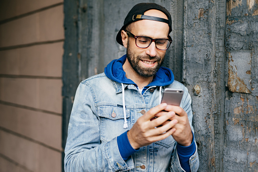 Horizontal portrait of handsome bearded stylish man in denim anorak wearing big eyeglasses holding smartphone using free Internet, checking e-mail, watching funny videos, updating applications