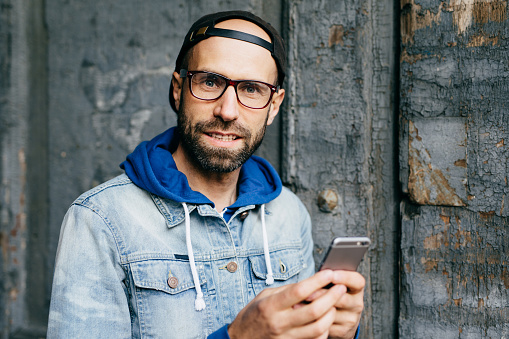 Close-up portrait of bearded blue-eyed man in stylish cap and denim anorak holding mobile phone surfing the Internet cheking his emails and communicating with friends. Technology and communication