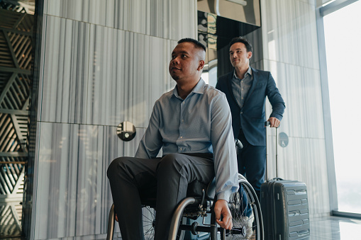 Two Malay businessmen on business travel one of whom is disabled wheeling their luggage out of the elevator lobby