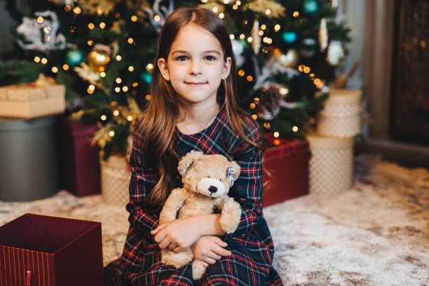 istock Small female child with appealing appearance, holds teddy bear, recieves present from parents, sits near Christmas tree. Adorable girl being glad to get gift on New Year. Christmas time. 1495574000
