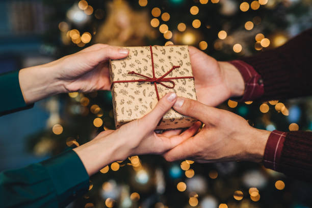 istock Wrapped box in woman`s and man`s hands. Affectionate husband congratulates his wife with New Year, presents her holiday gift box, stand against decorated Christmas tree. Holiday concept 1495574028