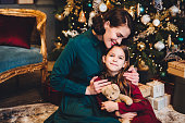 Affectionate mother embraces her little daughter as sit together near decorated Christmas tree. Small adorable girl being glad to recieve New Year present from her mom. Relationship concept.
