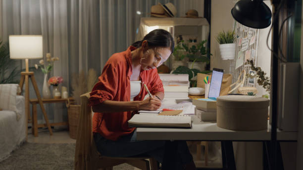 istock Young Asian woman use mask sheet on face sit front of desk with computer laptop and think create on work idea in living room at house at night. Work from home. 1495581948