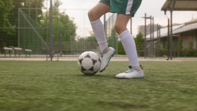 The boy dressed in soccer uniform is practicing on a mini football stadium. The young footballer skillfully maneuvers the ball, using feints, as he advances towards the goal. close up legs