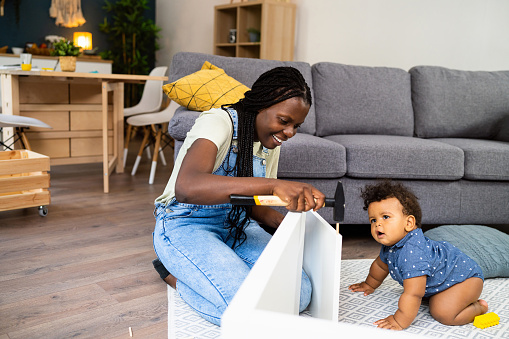Woman of black ethnicity assembling new furniture while her baby boy playing next to her.
