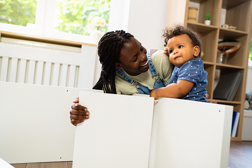 Woman of black ethnicity assembling piece of furniture while her curious baby boy makes her a company.