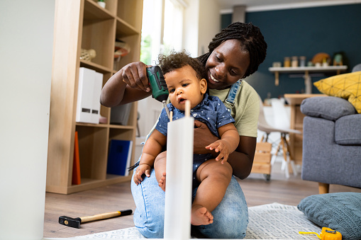 Woman of black ethnicity holding her baby boy while assembling new furniture.