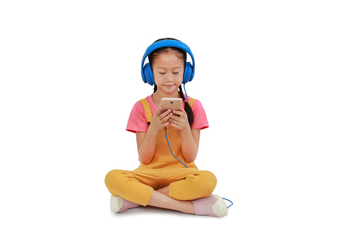 Asian little girl child wearing headphones and enjoy with smartphone sitting isolated on white background. Image with Clipping path