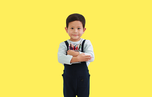 Portrait Asian little boy age 4 year old hugging a basket of color pencils isolated on yellow background. Image with Clipping path
