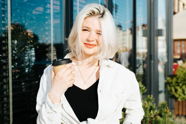 Charismatic cute blonde teenager with disposable cup of coffee posing near shop window on street looking at camera, squinting on sunny day. Pretty caucasian young woman outdoors, lifestyle stock photo