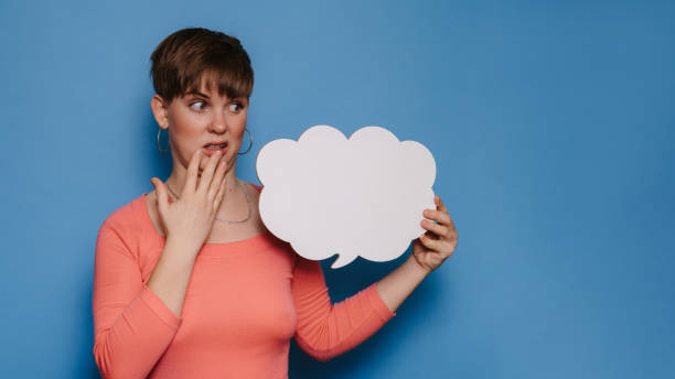 istock Studio shot of a young woman with a puzzled expression, holding a white empty banner in the form of a cloud on a blue background. Copy space, space for your ad or text. 1495592450
