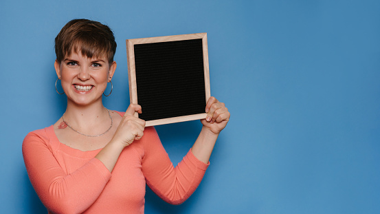 Studio shot of a smiling young woman holding a blank letter board on a blue background. Copy space, space for your ad or text