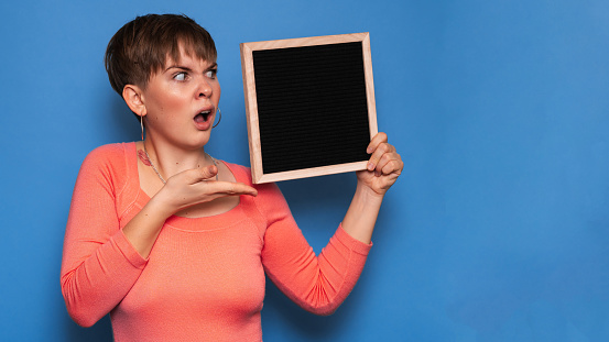 Studio shot of a young woman with a puzzled expression, holding a blank letter board on a blue background. Copy space, space for your ad or text