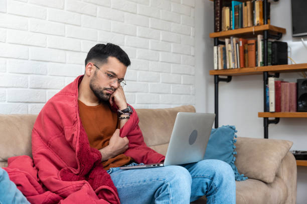 junger kranker, kranker, psychisch depressiver, ängstlicher mann, der allein zu hause sitzt, in eine decke gewickelt, und traurige filme auf einem laptop ansieht. verzweiflung, junger mann, gestresst, nachdem die beziehung untröstlich ausgebrochen ist - mentally ill stock-fotos und bilder