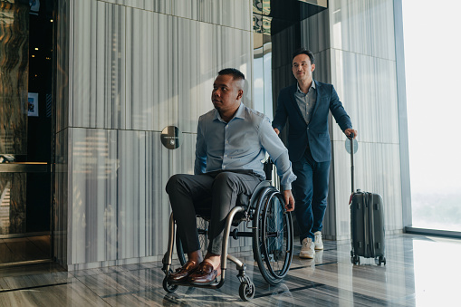 Two Malay businessmen on business travel one of whom is disabled wheeling their luggage out of the elevator lobby