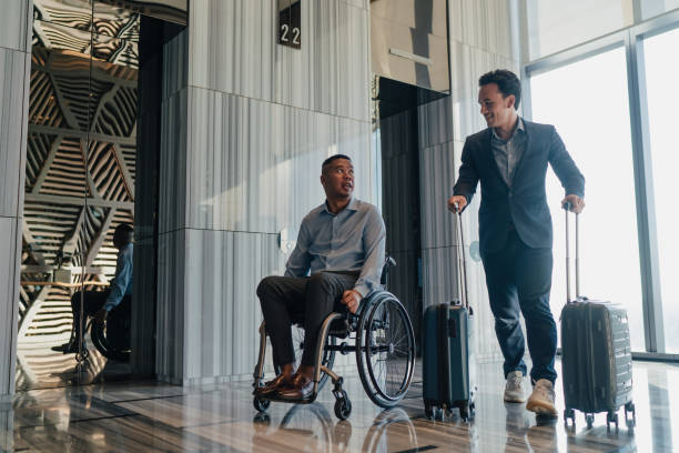 Two Malay businessmen on business travel one of whom is disabled wheeling their luggage out of the elevator lobby stock photo