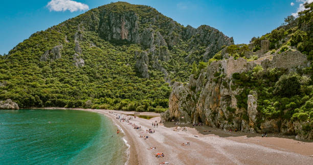 Steeped in history, Olympos Beach on the Lycian coast near Çiralı is especially popular with nature lovers.
Located in the middle of a nature reserve, the pebble beach will impress you with beautiful vegetation, ancient ruins, and a unique atmosphere.
The Olympos beach is situated just after the famous Cirali beach. The golden sandy Olympos beach is about 400 meters long, very beautiful and ideal for swimming and sunbathing.
With its pine forests and Beydaglari mountains at the background, wonderful nature, unique beach and sea, the historical site and ruins, Olympos is one of the most popular sights in Kemer. vista aérea de la playa de olympos en antalya, vista aérea de la playa de cirali en el mar mediterráneo en la provincia de antalya, naturaleza y escena de relajación del mar, escena de verano colorida de fondo, popular destino de vacaciones de turquía
