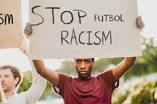Serious young man in casual clothes holding Stop Racism sign looking at the camera while protesting standing on the street.