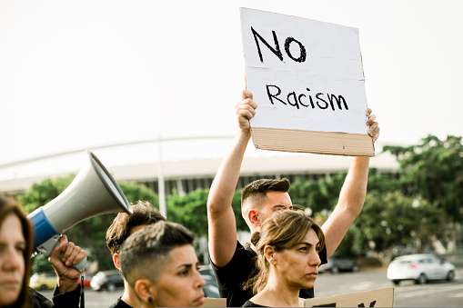 From below a group of young people in casual clothes standing with their arm raised and holding a sign with the inscription No Racism.
