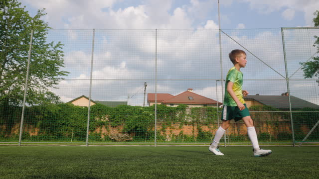 The boy dressed in soccer uniform is practicing on a mini football stadium. The young footballer kicks the ball towards the goal. Profile full shot