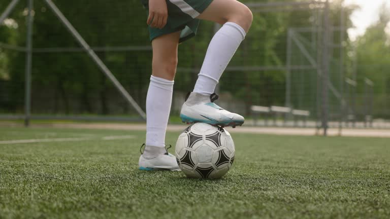 The boy dressed in soccer uniform is practicing on a mini football stadium. A close-up shot of the legs of the young footballer, with one foot is on the ball