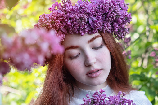 young girl with long red hair with a wreath of lilacs on her head in the garden in spring