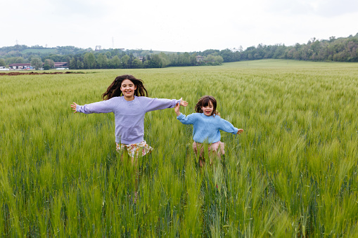 two girls in jumpers running together in a big field of green wheat