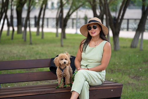 An Asian girl in sunglasses and a hat is sitting on a bench with her dog (that poodle). Summer portrait of a young woman outdoors.