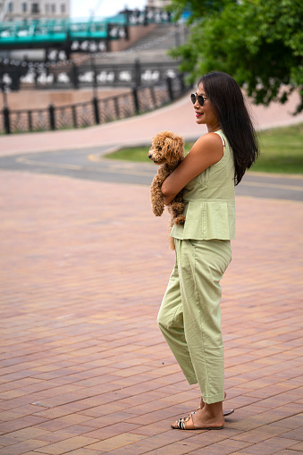 An Asian girl in sunglasses walks along the promenade with her dog (that poodle). Summer portrait of a young woman outdoors.
