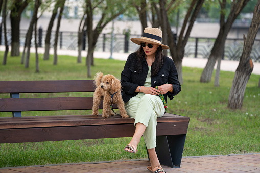 An Asian girl in sunglasses and a hat is sitting on a bench with her dog (that poodle). Summer portrait of a young woman outdoors.