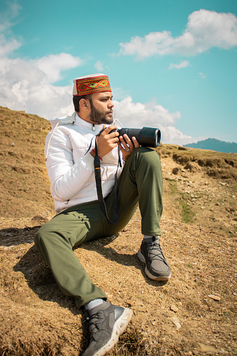 Young traveler man sitting on top of the mountain and taking a picture of a beautiful landscape of Bir billing mountain hills with dslr camera.