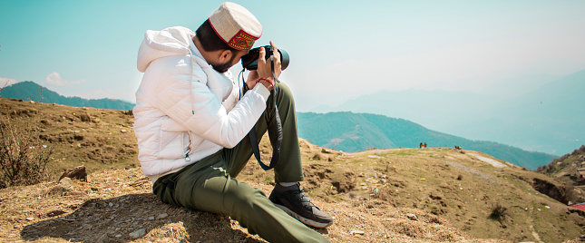 Young traveler man sitting on top of the mountain and taking a picture of a beautiful landscape of Bir billing mountain hills with dslr camera. Panoramic image with copy space.