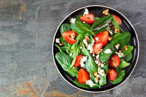 Summer salad of spinach, strawberries and blue cheese in a black plate. Overhead view on a dark slate background.