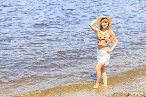 A Japanese woman wading in Burrard Inlet, Vancouver BC. She is wearing long blond wavy hair, makeup, a straw hat, a brown sleeveless top, blue mini skirt and barefoot.