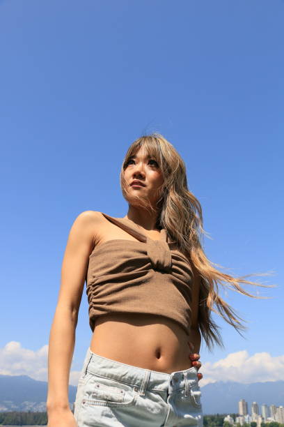 Windblown Woman A Japanese woman on a sunny windy day with Vancouver BC in the background with Stanley Park. She is wearing long, blond wavy tousled hair, makeup, a brown sleeveless crop top and blue mini skirt. crop top view stock pictures, royalty-free photos & images