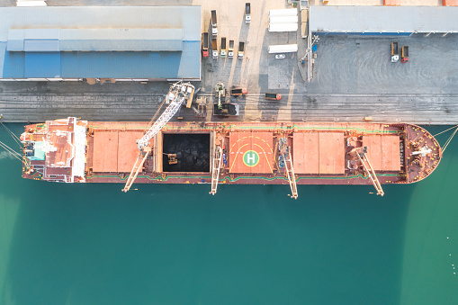 The tug accompanies the ship when entering the cargo port. \nAerial drone view above sea port. Cargo vessel is loading containers and bulk. Professional business logistics and transportation of cargo ship.