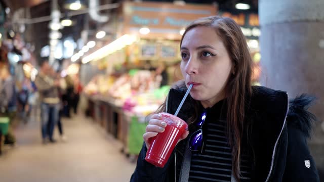 Side view of latin young woman in outerwear drinking fresh juice from plastic cup while visiting dessert stall in Santa Caterina market in Barcelona, Spain
