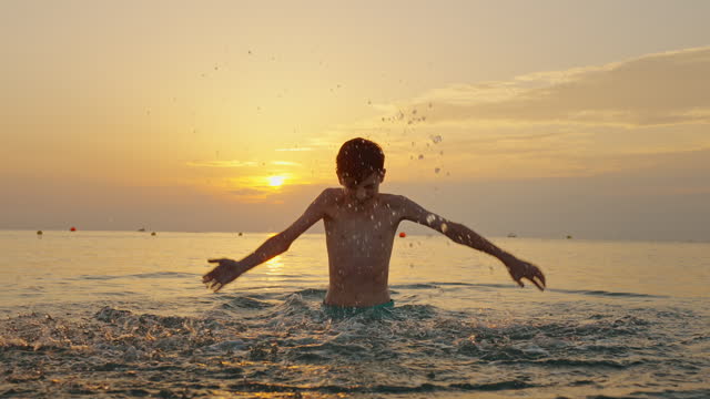 Happy male runs jumps merrily splashing in sea water in summer on warm disk sun sunset slow motion. Large splashes of water close up. Child plays on seashore. Childhood. Go Everywhere. Travel