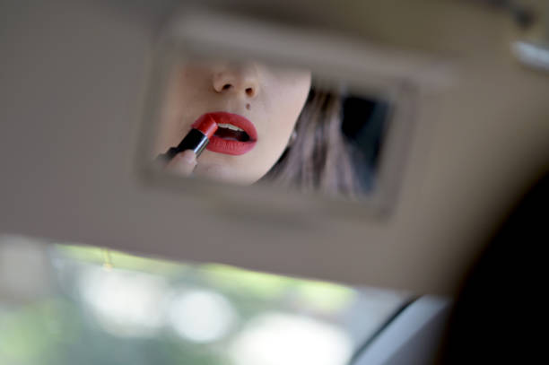 Distracted driver. Young attractive woman looking in rear view mirror painting her lips doing applying make up while driving the car.