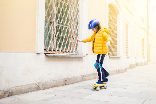 a boy in a yellow jacket, with blue protection on his arms and legs, with a purple helmet with a plastic skateboard in the city in cold weather