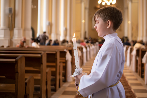 the boy holds a burning candle in his hands, stands in white clothes in the temple before the children's mass